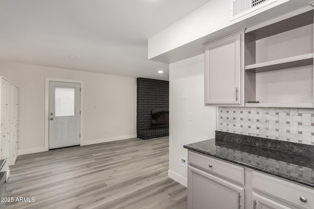 kitchen featuring open shelves, light wood-style flooring, decorative backsplash, a brick fireplace, and dark stone countertops