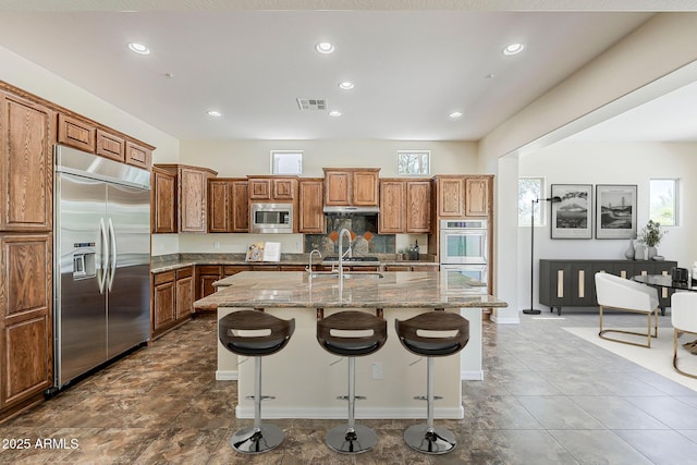 30374 N 124th Dr, Peoria, AZ 85383 - kitchen featuring built in appliances, a kitchen breakfast bar, brown cabinets, light stone counters, and an island with sink