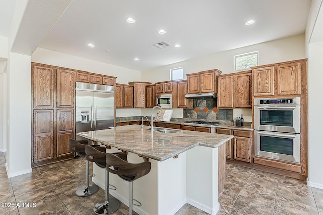 30374 N 124th Dr, Peoria, AZ 85383 - kitchen featuring built in appliances, a sink, a center island with sink, light stone countertops, and brown cabinetry