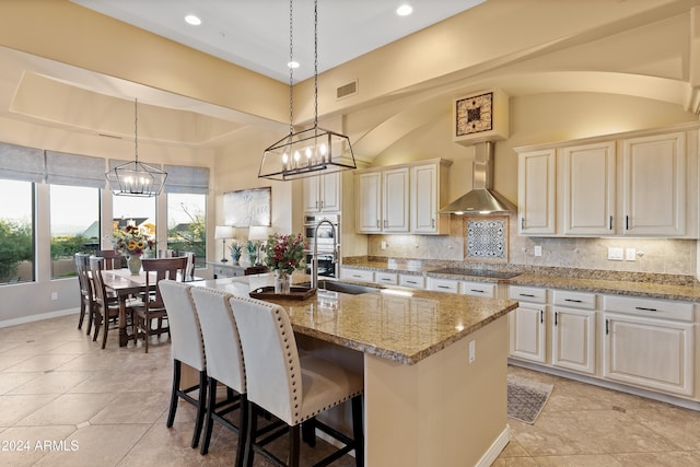 kitchen with hanging light fixtures, wall chimney exhaust hood, decorative backsplash, light stone countertops, and an island with sink