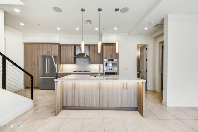 kitchen with a center island with sink, appliances with stainless steel finishes, pendant lighting, decorative backsplash, and wall chimney range hood