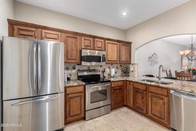 kitchen with light stone counters, stainless steel appliances, a sink, brown cabinetry, and stone tile flooring