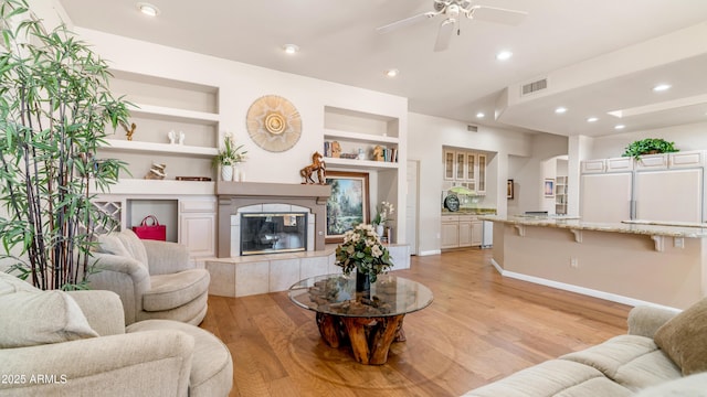 living area with light wood-type flooring, a tiled fireplace, visible vents, built in features, and ceiling fan