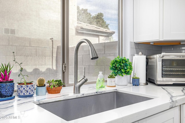 kitchen view of a sink, white cabinetry, light stone counters, and backsplash