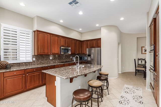 kitchen with an island with sink, backsplash, a kitchen breakfast bar, stainless steel appliances, and dark stone countertops