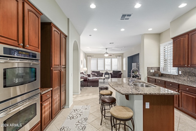 kitchen featuring stainless steel double oven, a kitchen island with sink, sink, a breakfast bar, and ceiling fan