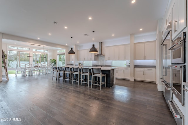 kitchen with wall chimney exhaust hood, white cabinets, pendant lighting, a kitchen island with sink, and backsplash