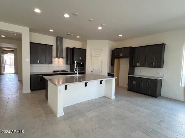 kitchen featuring wall chimney range hood, gas cooktop, backsplash, double oven, and an island with sink