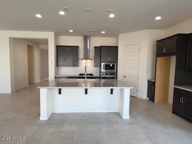 kitchen with a breakfast bar, backsplash, stainless steel appliances, a center island with sink, and wall chimney exhaust hood