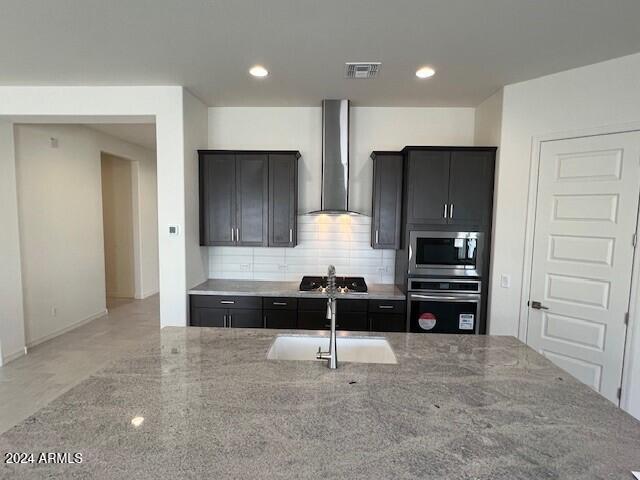 kitchen featuring sink, tasteful backsplash, light stone counters, stainless steel appliances, and wall chimney range hood