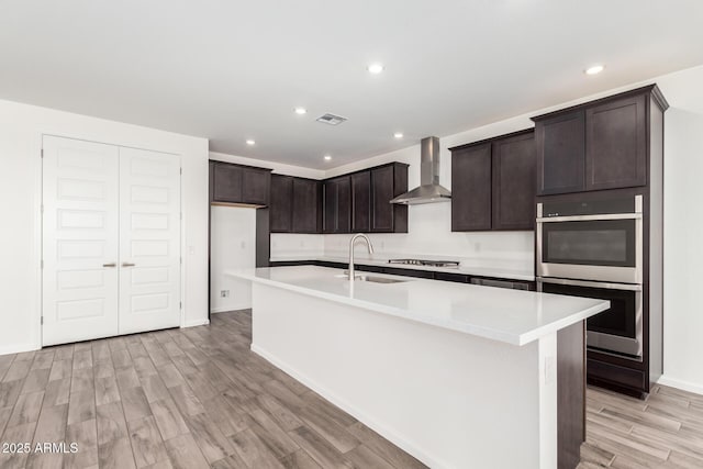 kitchen featuring stainless steel appliances, a center island with sink, light hardwood / wood-style floors, and wall chimney exhaust hood