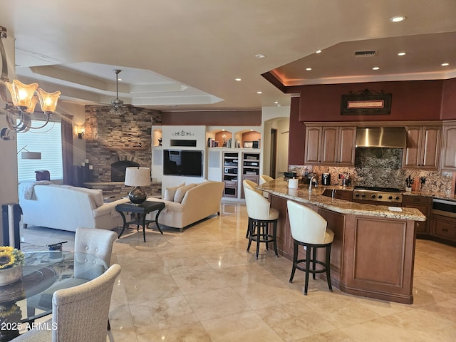 kitchen with a tray ceiling, a kitchen breakfast bar, light stone countertops, and wall chimney range hood