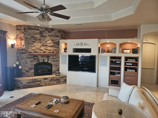 living room featuring ceiling fan, a stone fireplace, a raised ceiling, and light tile patterned floors