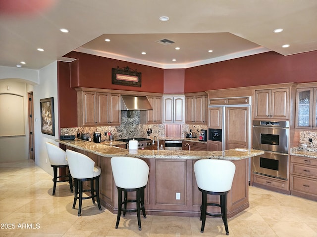 kitchen featuring light stone counters, stainless steel double oven, a kitchen breakfast bar, and wall chimney range hood