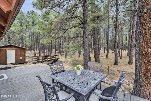 wooden terrace with an outdoor structure, outdoor dining space, a forest view, and a storage shed