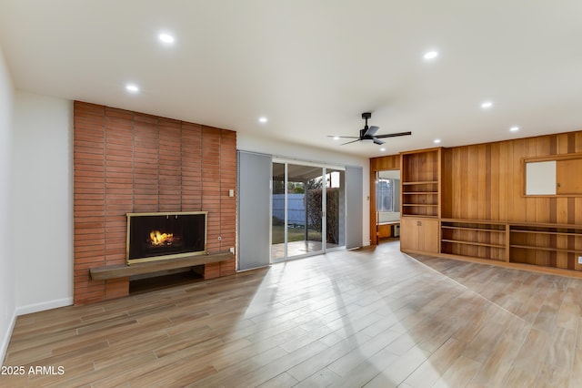 unfurnished living room with ceiling fan, a fireplace, wood walls, and light wood-type flooring