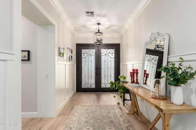foyer entrance featuring light wood finished floors, french doors, wainscoting, a decorative wall, and crown molding