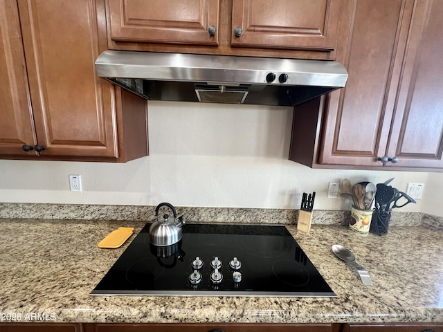 kitchen featuring light stone countertops, wall chimney exhaust hood, black electric cooktop, and brown cabinetry