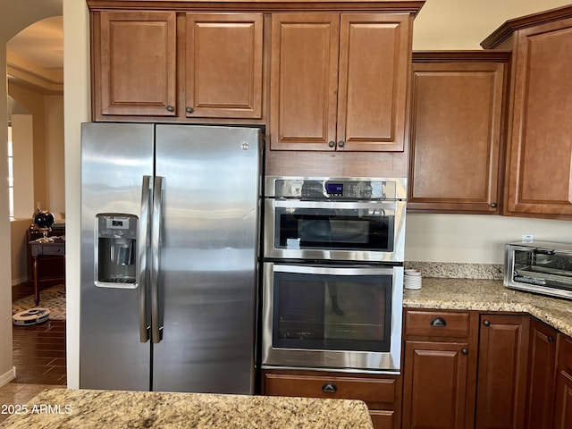 kitchen with a toaster, arched walkways, appliances with stainless steel finishes, brown cabinets, and light stone countertops