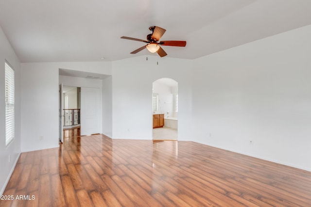 empty room featuring a ceiling fan, wood finished floors, arched walkways, baseboards, and vaulted ceiling