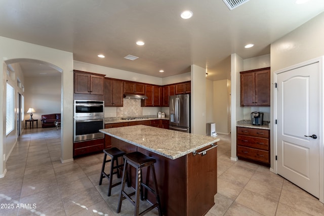 kitchen featuring a center island, under cabinet range hood, a breakfast bar, arched walkways, and appliances with stainless steel finishes