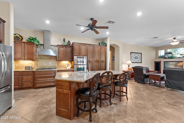 kitchen featuring light stone countertops, wall chimney range hood, a kitchen island with sink, a breakfast bar, and appliances with stainless steel finishes