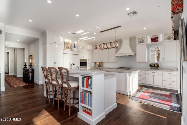 kitchen featuring custom exhaust hood, hanging light fixtures, paneled refrigerator, a kitchen island with sink, and white cabinets