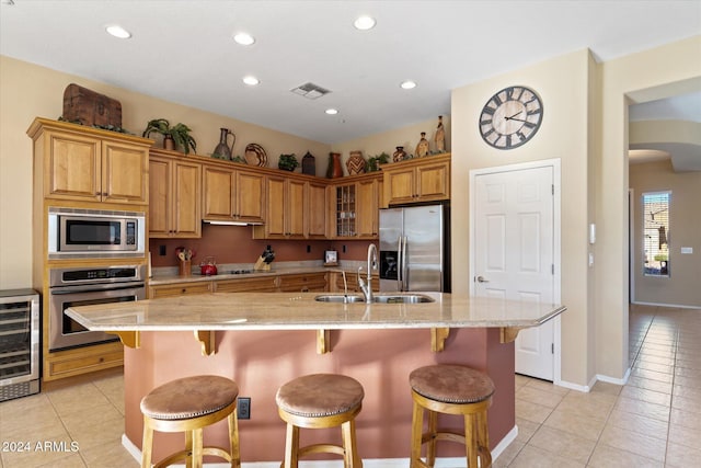 kitchen with stainless steel appliances, sink, light tile patterned floors, wine cooler, and an island with sink