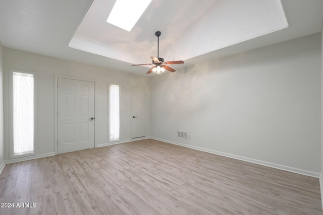 empty room with a tray ceiling, a skylight, ceiling fan, and light hardwood / wood-style floors