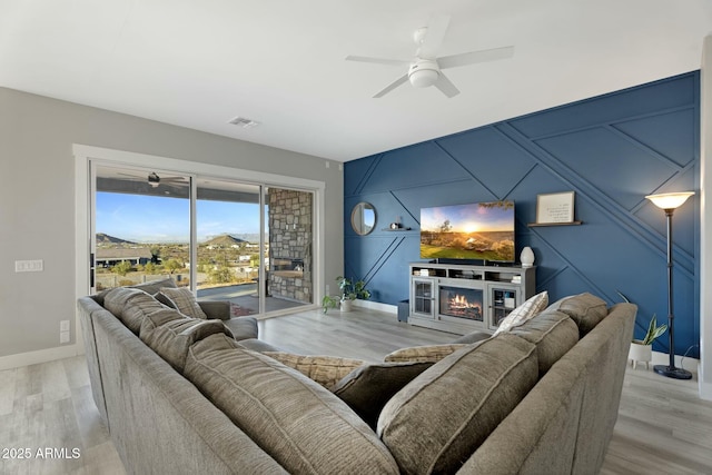 living area featuring an accent wall, a ceiling fan, baseboards, light wood-type flooring, and a glass covered fireplace