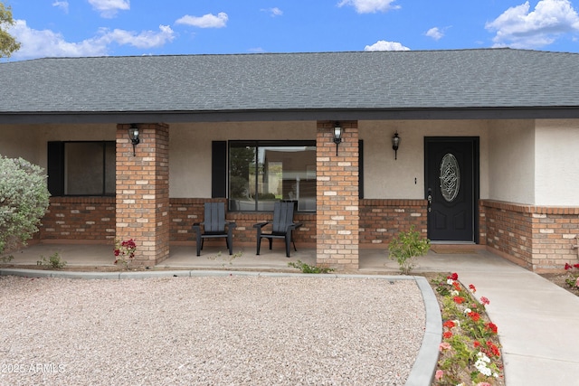 doorway to property with brick siding, covered porch, roof with shingles, and stucco siding