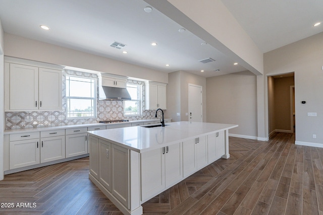 kitchen featuring stainless steel gas cooktop, a center island with sink, backsplash, a sink, and under cabinet range hood