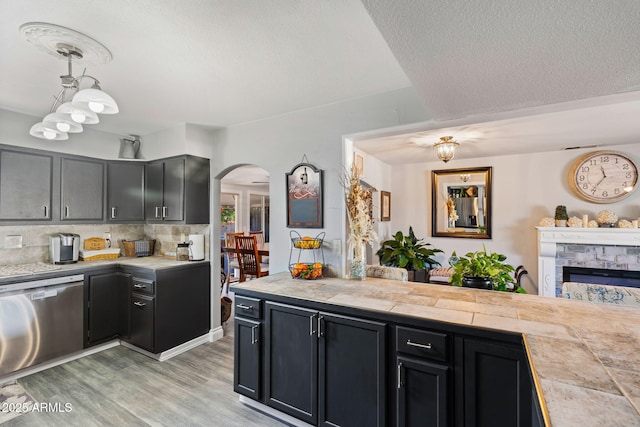 kitchen with dishwasher, hanging light fixtures, light wood-type flooring, a stone fireplace, and tile countertops