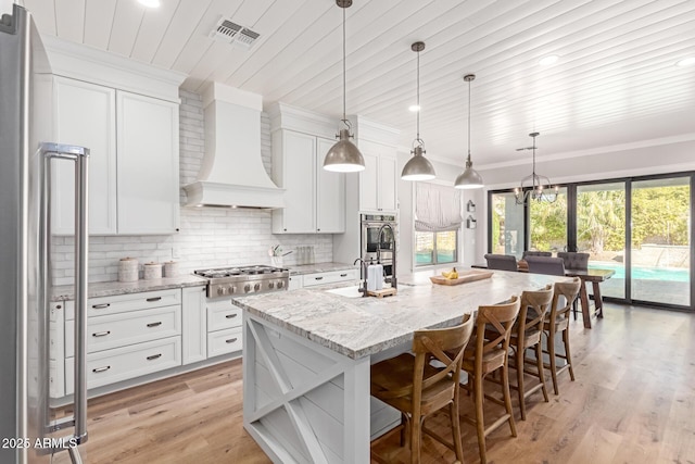 kitchen with appliances with stainless steel finishes, a kitchen island with sink, hanging light fixtures, white cabinets, and custom exhaust hood