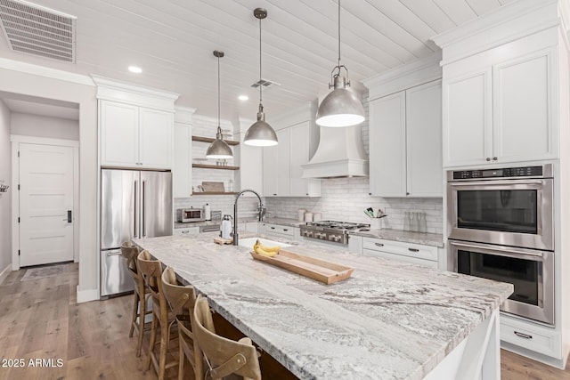 kitchen featuring pendant lighting, premium range hood, a kitchen island with sink, stainless steel appliances, and white cabinets