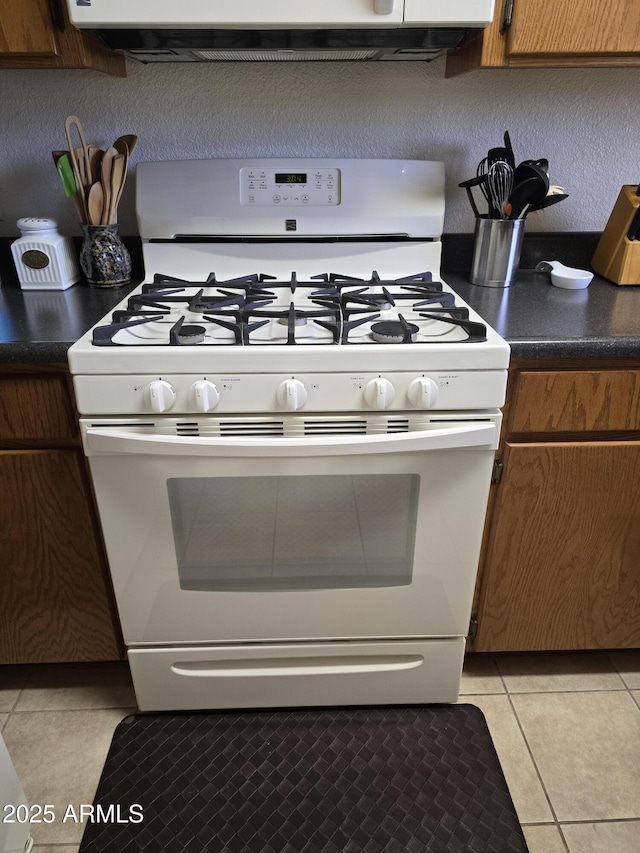 kitchen featuring brown cabinetry, dark countertops, white range with gas stovetop, and light tile patterned floors