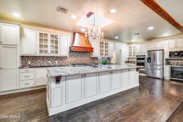 kitchen featuring appliances with stainless steel finishes, light stone counters, custom exhaust hood, a kitchen island with sink, and pendant lighting