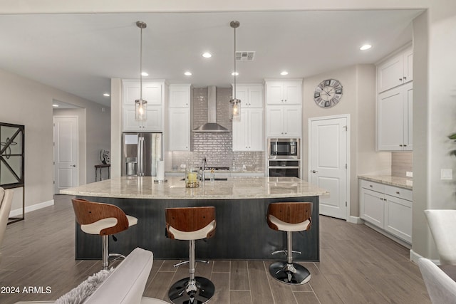 kitchen featuring appliances with stainless steel finishes, wall chimney range hood, wood-type flooring, white cabinets, and hanging light fixtures