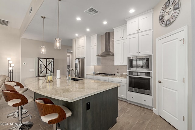 kitchen featuring white cabinets, wall chimney range hood, hanging light fixtures, an island with sink, and stainless steel appliances