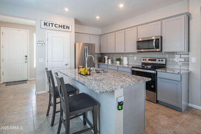 kitchen featuring appliances with stainless steel finishes, a center island with sink, light stone counters, decorative backsplash, and gray cabinets