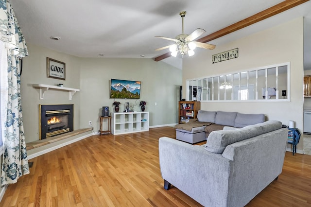 living room featuring a ceiling fan, lofted ceiling with beams, wood finished floors, a glass covered fireplace, and baseboards