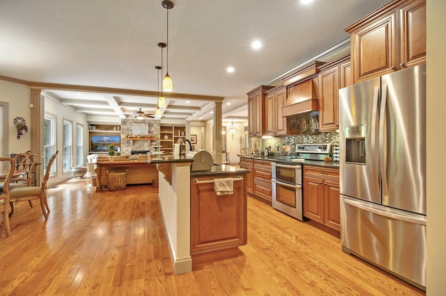 kitchen with brown cabinetry, an island with sink, appliances with stainless steel finishes, decorative light fixtures, and premium range hood