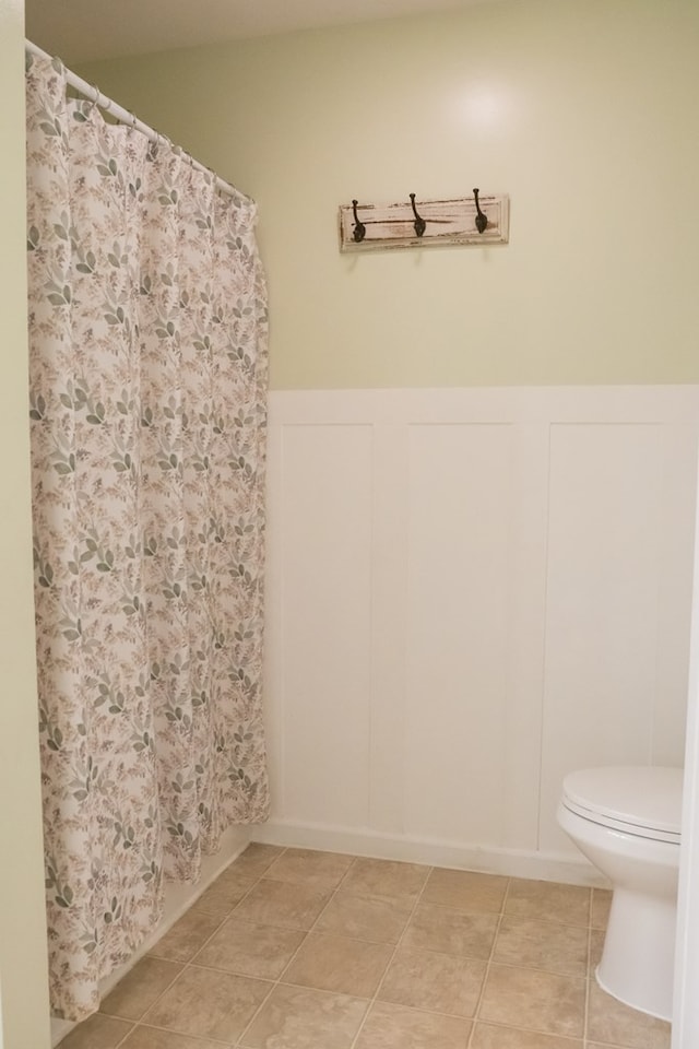 full bath featuring tile patterned flooring, a decorative wall, toilet, and a wainscoted wall