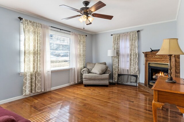 living area with ornamental molding, ceiling fan, wood finished floors, a warm lit fireplace, and baseboards