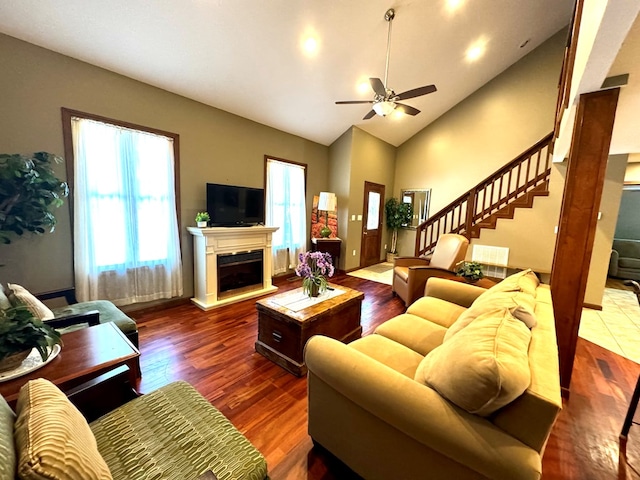 living room with a fireplace with raised hearth, high vaulted ceiling, wood finished floors, a ceiling fan, and stairs