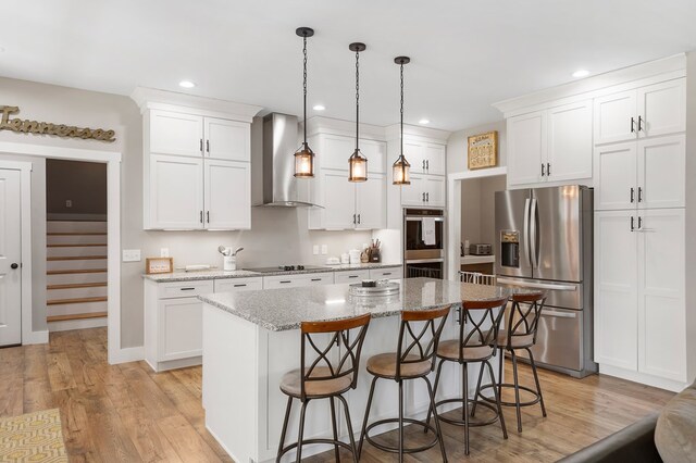 kitchen with white cabinets, appliances with stainless steel finishes, light stone counters, a center island, and wall chimney range hood