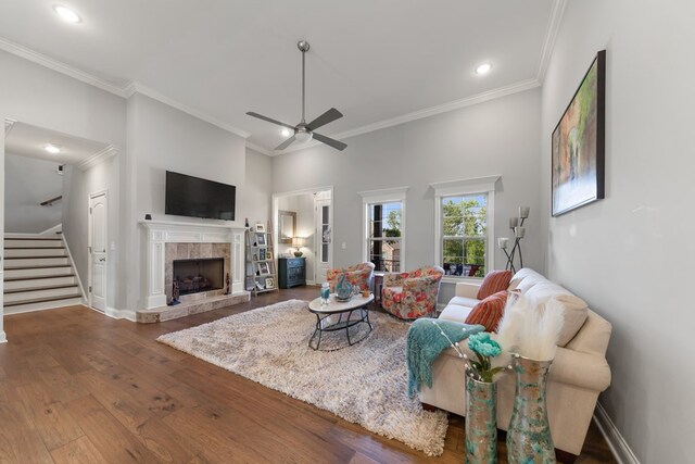 living room featuring a tiled fireplace, ceiling fan, wood finished floors, stairs, and crown molding