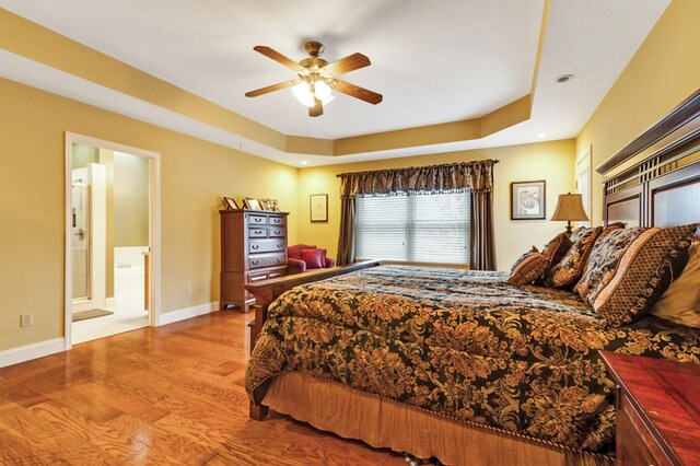 bedroom with ensuite bathroom, ceiling fan, wood finished floors, baseboards, and a tray ceiling