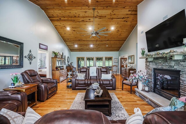 living area with a towering ceiling, wood ceiling, ceiling fan, a stone fireplace, and light wood-type flooring