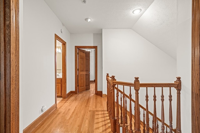 hall featuring baseboards, vaulted ceiling, a textured ceiling, an upstairs landing, and light wood-type flooring